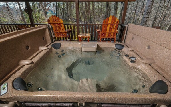 Un jacuzzi con dos sillas Adirondack y una mesa pequeña, rodeado por una terraza de madera con vistas a un bosque.