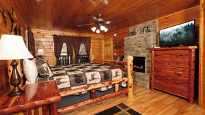 Ein gemütliches Blockhauszimmer mit Bett, Holzkommode, Kamin und Fernseher.