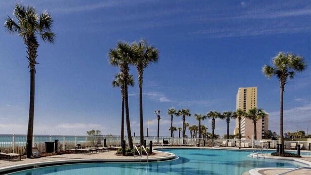 A large outdoor swimming pool with palm trees, a clear blue sky, and a building in the background.