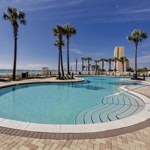 A large outdoor swimming pool with palm trees, a clear blue sky, and a building in the background.