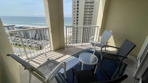 A balcony with a view of the ocean and a parking lot. There are two chairs and a small table.