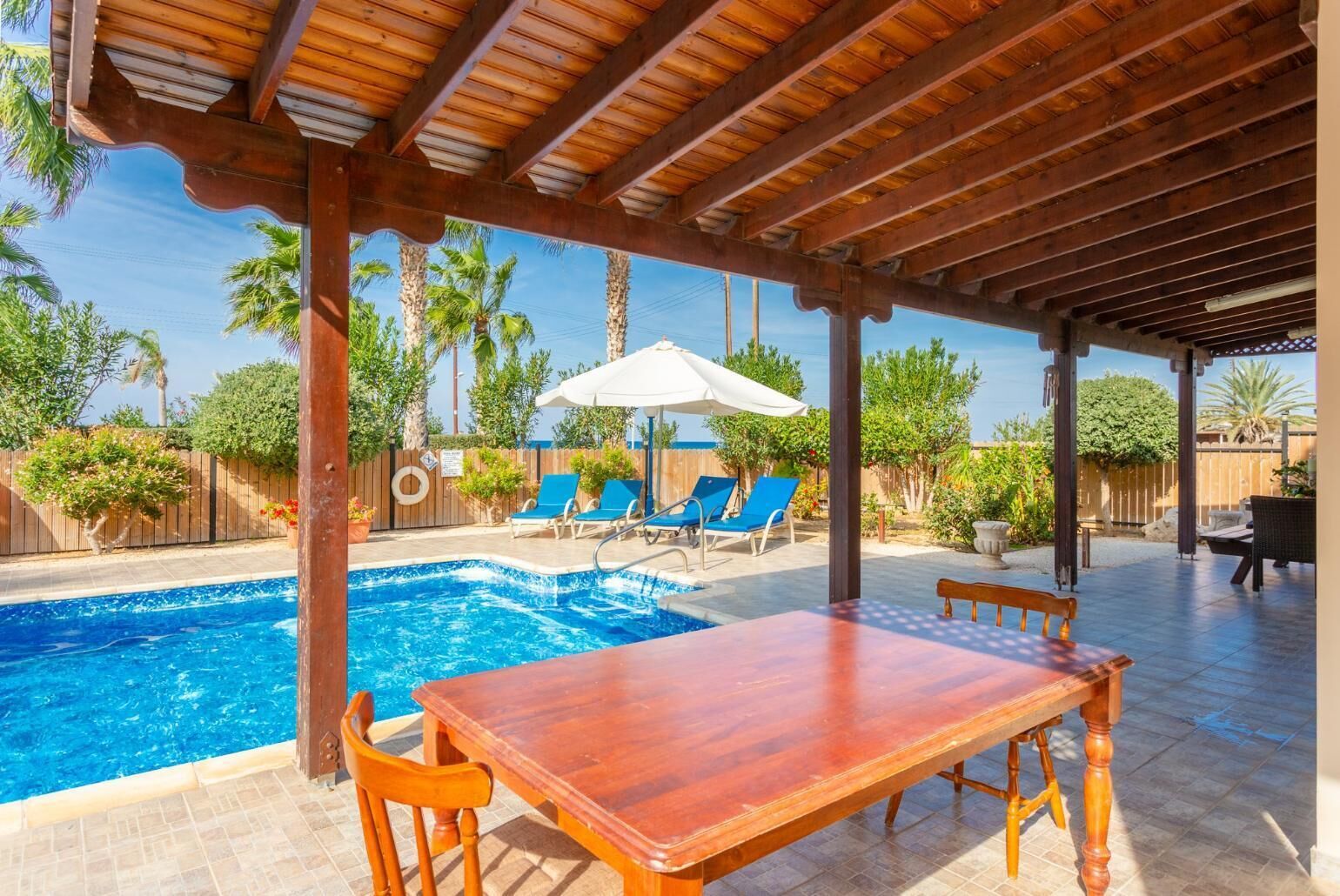 A covered outdoor pool area with a wooden table and chairs, lounge chairs, and a white umbrella.