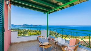 A balcony with a view of the sea, two wicker chairs, a table with a bottle and glasses, and a green railing.