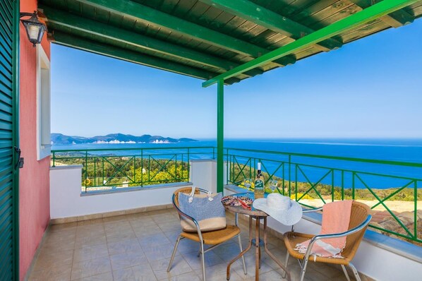 Un balcon donnant sur la mer, deux fauteuils en osier, une table avec une bouteille et des verres, et une rambarde verte.