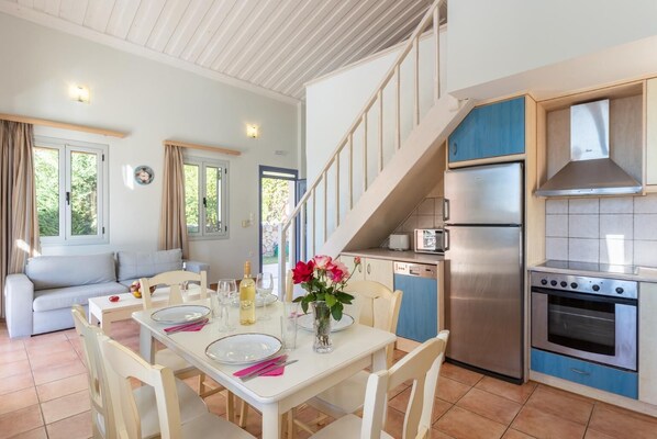 Una cocina moderna con horno empotrado, lavavajillas y refrigerador de acero inoxidable. Hay un área de comedor con una mesa preparada para dos personas, y una escalera que conduce a un nivel superior.