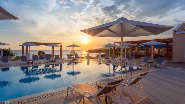 Un espace piscine doté de chaises longues et de parasols, offrant une vue sur une étendue d’eau au coucher du soleil.