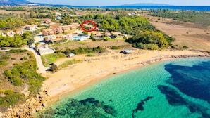 An aerial view of a coastal area with a beach, clear blue waters, and a cluster of buildings with a swimming pool.