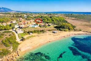Vista aérea de uma área costeira com praia, águas azul-claras e um conjunto de edifícios com piscina.