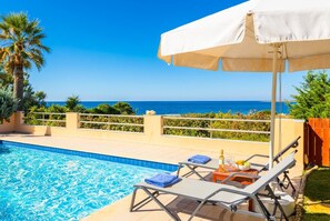 Uma área à beira da piscina com vista para o mar, uma espreguiçadeira, uma mesa com uma garrafa e copos, e um guarda-sol.