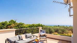 Un balcon avec un canapé, une chaise et une table où sont posées des boissons et un livre. Le balcon offre une vue sur un paysage verdoyant et la mer.