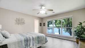 A bedroom with a bed, a ceiling fan, a large window with a view of trees and water, and a plant.