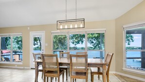 A dining area with a large table and six chairs, large windows with a view of trees and water, and a hanging light fixture.
