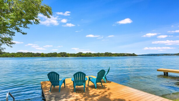 A wooden dock with three green Adirondack chairs facing a calm lake, surrounded by trees and a clear sky.