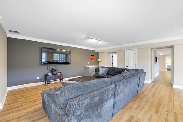 A modern living room with a large grey sectional sofa, a flat-screen TV mounted on the wall, and a wooden floor. - 4.9-Star Rated, Stunning 3600 SF House on Golf Course (Waleska)