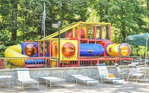 Un parque infantil con toboganes y estructuras para escalar, rodeado de árboles y una zona de piscina con sillas de descanso.