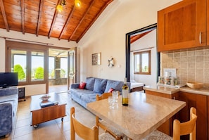 A spacious living area with a sofa, a coffee table, and a dining table with chairs. There is a television on a stand, a bottle of wine, and a bowl of fruit on the table. - Villa Katerina Private Pool, Sea Views, A/C, WiFi (Skopelos)