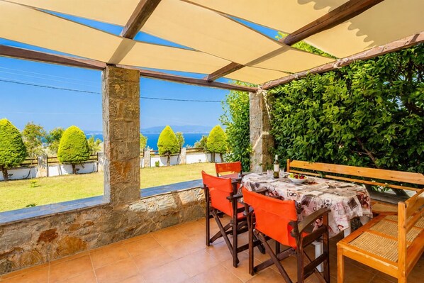 Un espace extérieur aménagé, abrité et doté de tables et de chaises, offrant une vue sur un jardin paysager et sur la mer.
