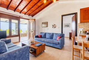 Un vaste espace de vie comprenant un canapé bleu, une table basse et un téléviseur. On y trouve également un coin repas avec une table et des chaises. La pièce est dotée de poutres apparentes en bois et de grandes fenêtres.
