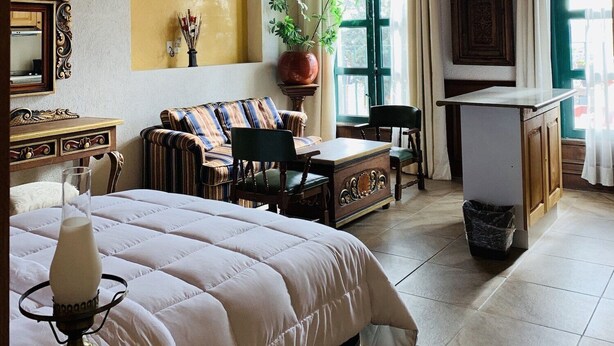 A room with a high, vaulted ceiling, a bed with a white duvet, a wooden desk, a chair, a small table, and a potted plant. - Posada la Ermita (San Miguel de Allende)