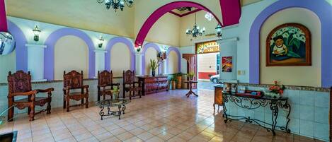 A spacious lobby with high ceilings, ornate wooden chairs, a reception desk, and a tiled floor.