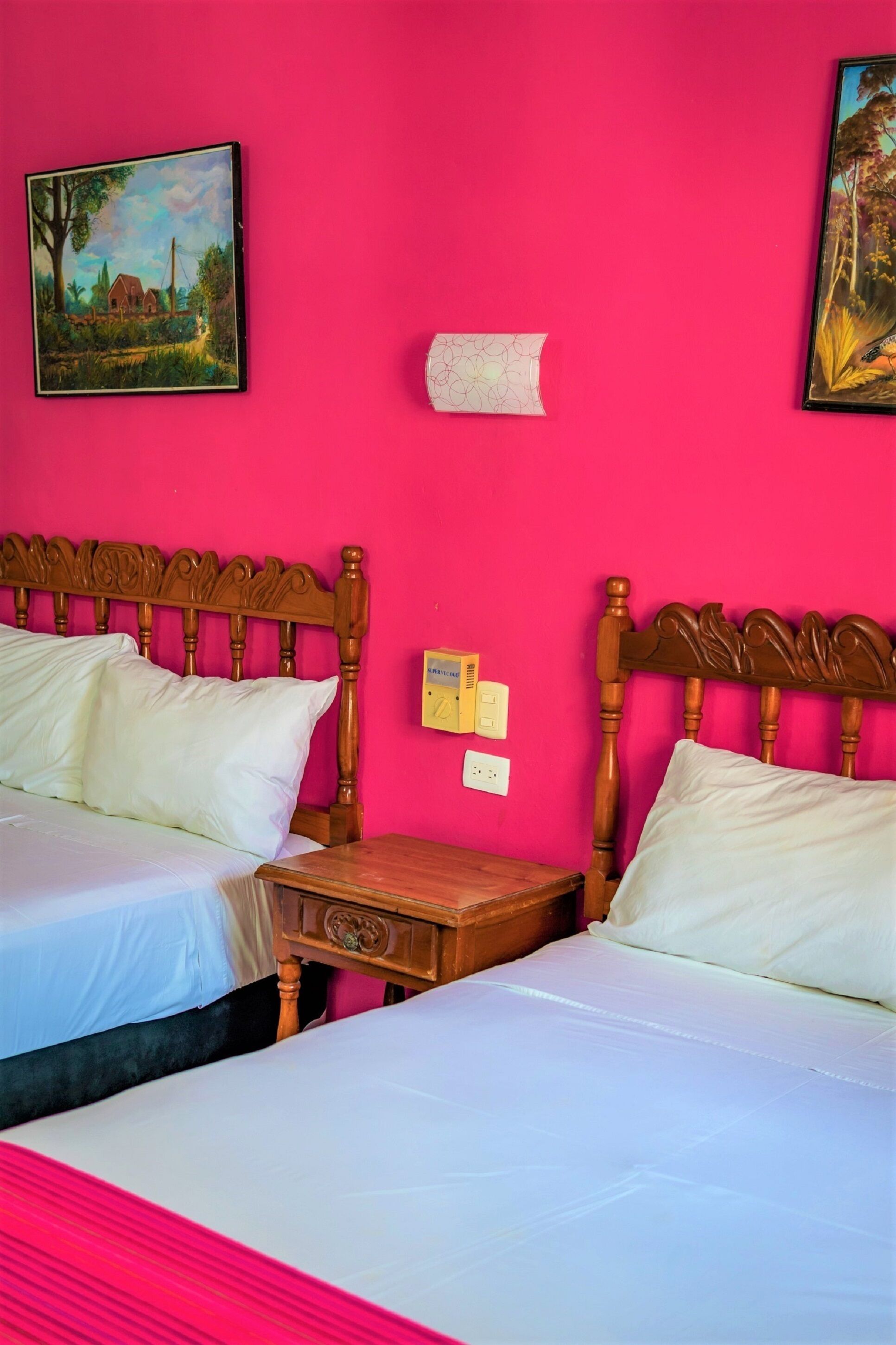 A room with two beds, wooden headboards, white bedding, a wooden nightstand, and a wall-mounted light switch.