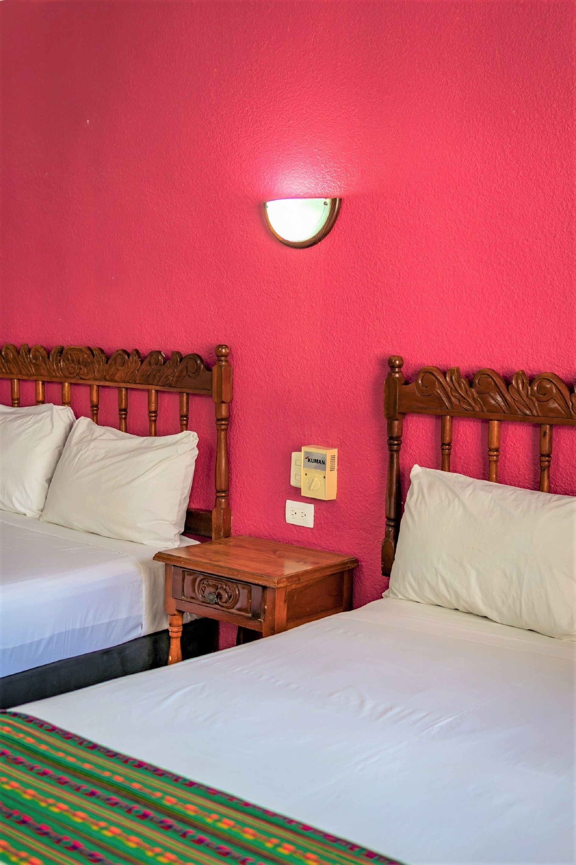 A room with two beds, a wooden nightstand, a wall-mounted light, and a thermostat.