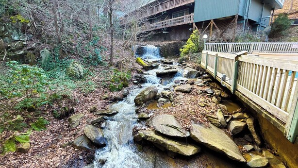 Un arroyo serpentea por un área boscosa, con un puente de madera y un edificio al fondo.