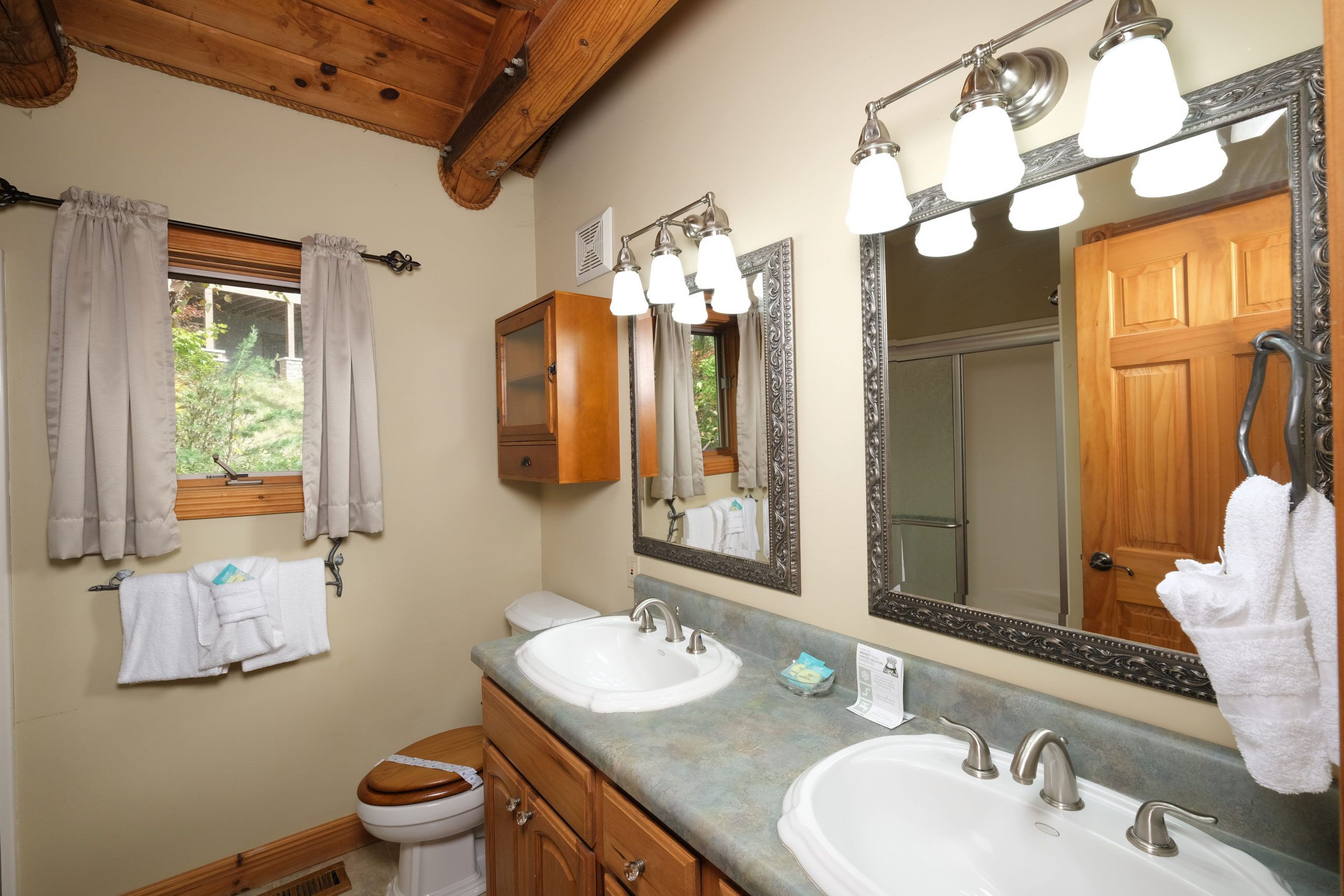 A bathroom with two sinks, a toilet, a window with curtains, and a large mirror with lights.