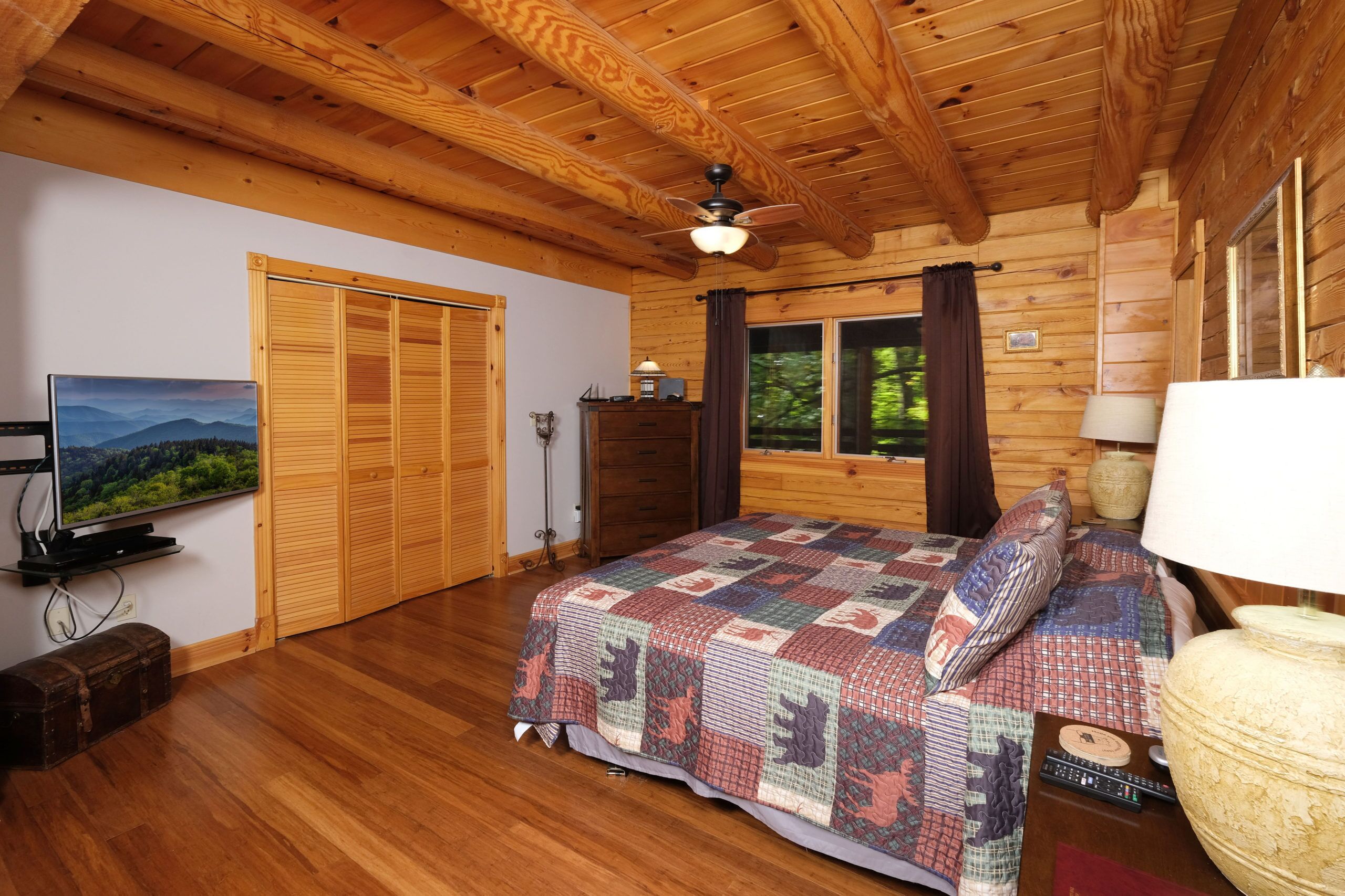 A wooden cabin bedroom with a bed, a television, a ceiling fan, and a window with curtains.