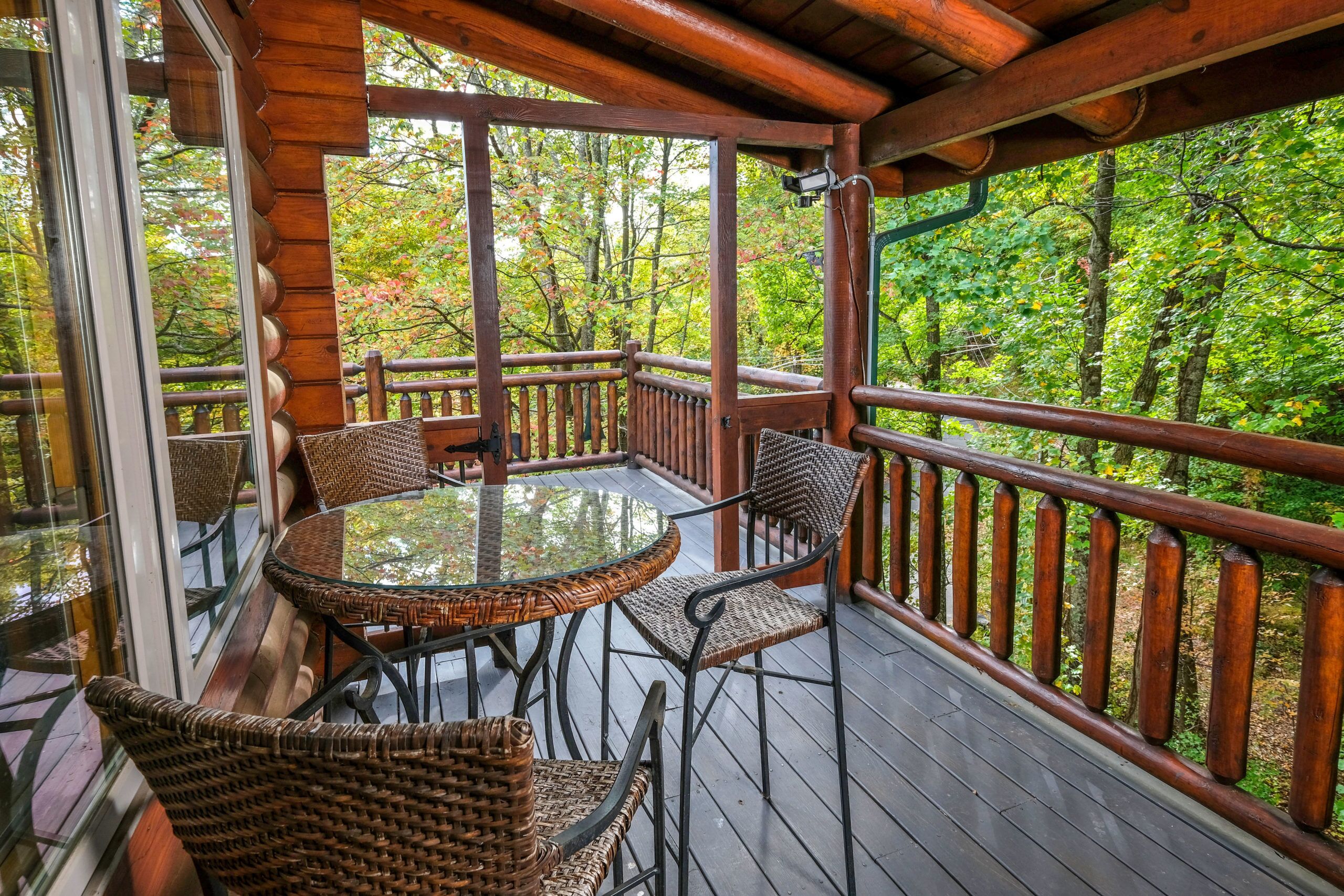 A wooden deck with a glass table and wicker chairs, surrounded by a wooden railing, overlooking a forested area.