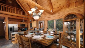 L'intérieur d'un chalet en bois comprend une salle à manger avec une table dressée pour quatre personnes, une cuisine équipée d'appareils électroménagers modernes et une grande fenêtre donnant sur un paysage verdoyant.