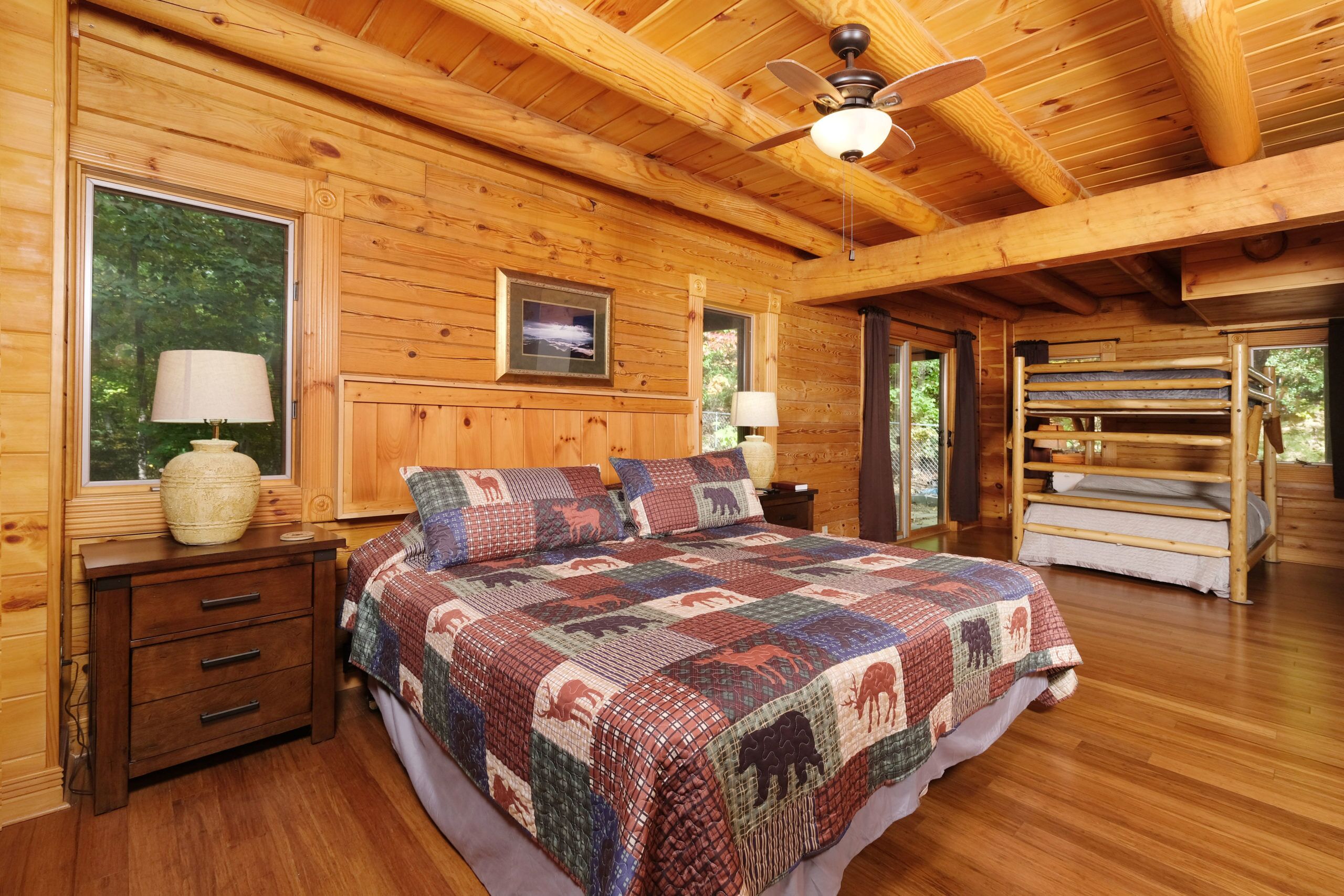 A wooden cabin bedroom with a large bed, two bedside tables, a ceiling fan, and a bunk bed in the corner.