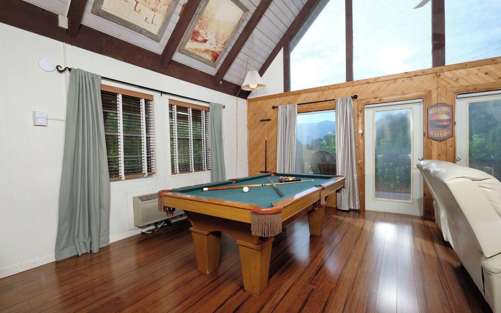 A pool table in a room with wooden floors, large windows, and a view of the outdoors.