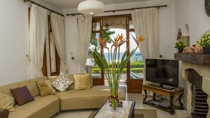 Olohuoneessa on beige sohva, sohvapöytä, televisio ja ikkunasta avautuu näkymä merelle. Ikkunassa on verhot.