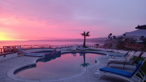 A pool area with lounge chairs and a palm tree, overlooking a body of water and a distant cityscape during sunset.