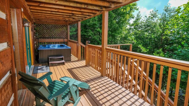 Una terraza de madera con un jacuzzi, sillas verdes y una barandilla de madera con vistas a un área boscosa.
