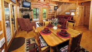 Un acogedor interior de cabaña con una mesa de comedor dispuesta para cuatro, una chimenea, un televisor montado en la pared y una escalera que conduce a otro nivel.