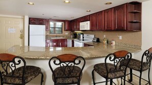 A kitchen with granite countertops, dark wooden cabinets, a white refrigerator, a microwave, a stove, and a sink. There are four bar stools with floral upholstery. - Creek View GH403 - Sip coffee on the balcony with the sound of the creek! (Gatlinburg)