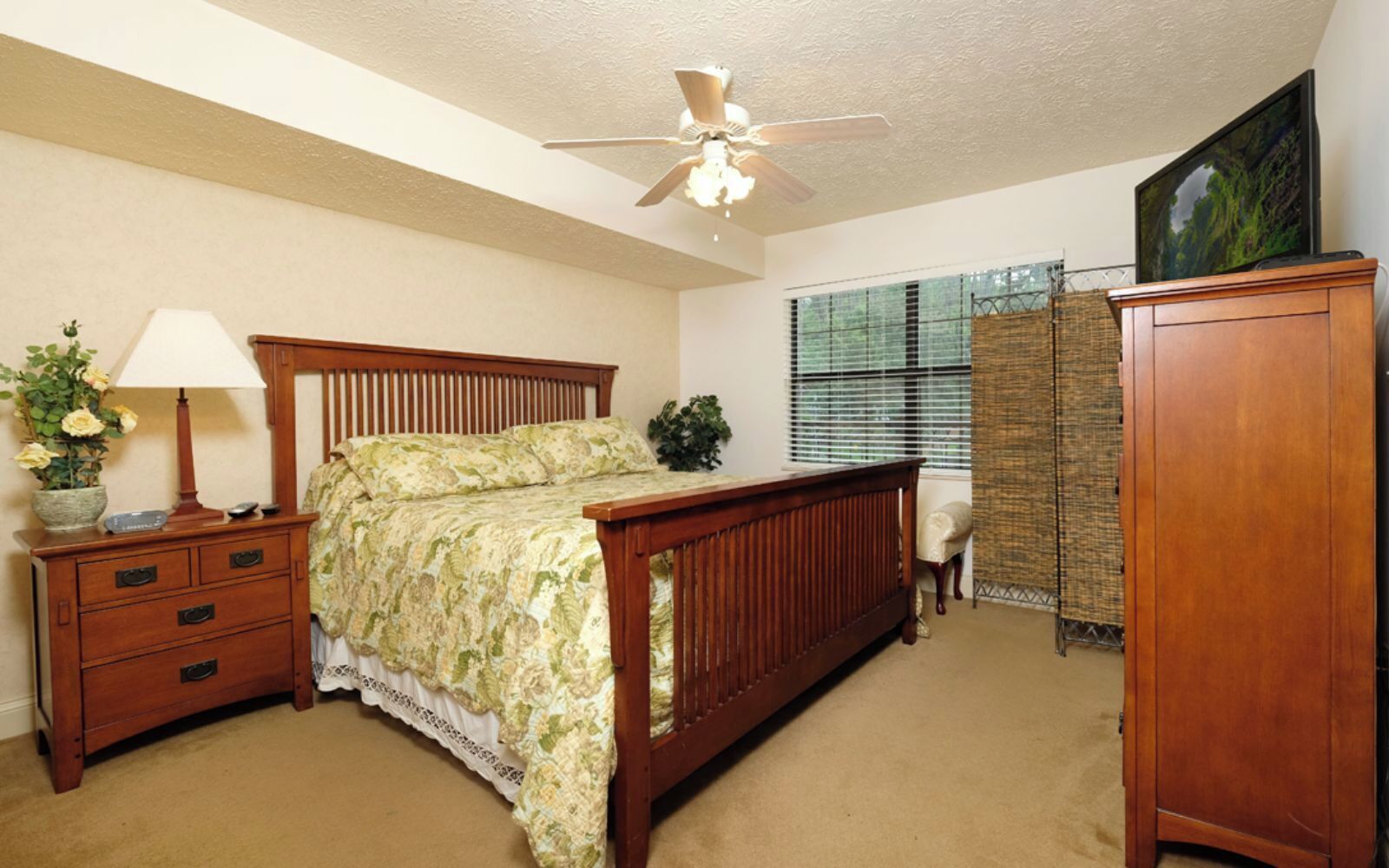 A bedroom with a wooden bed, a nightstand, a lamp, a dresser, and a window with blinds.