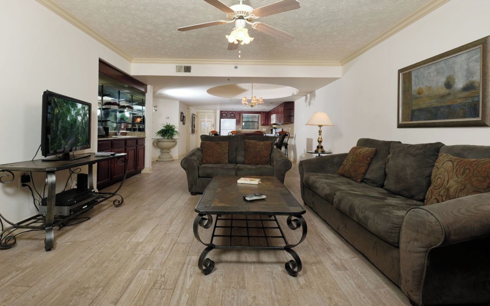 A living room with a television, a coffee table, two sofas, a ceiling fan, and a kitchen in the background.