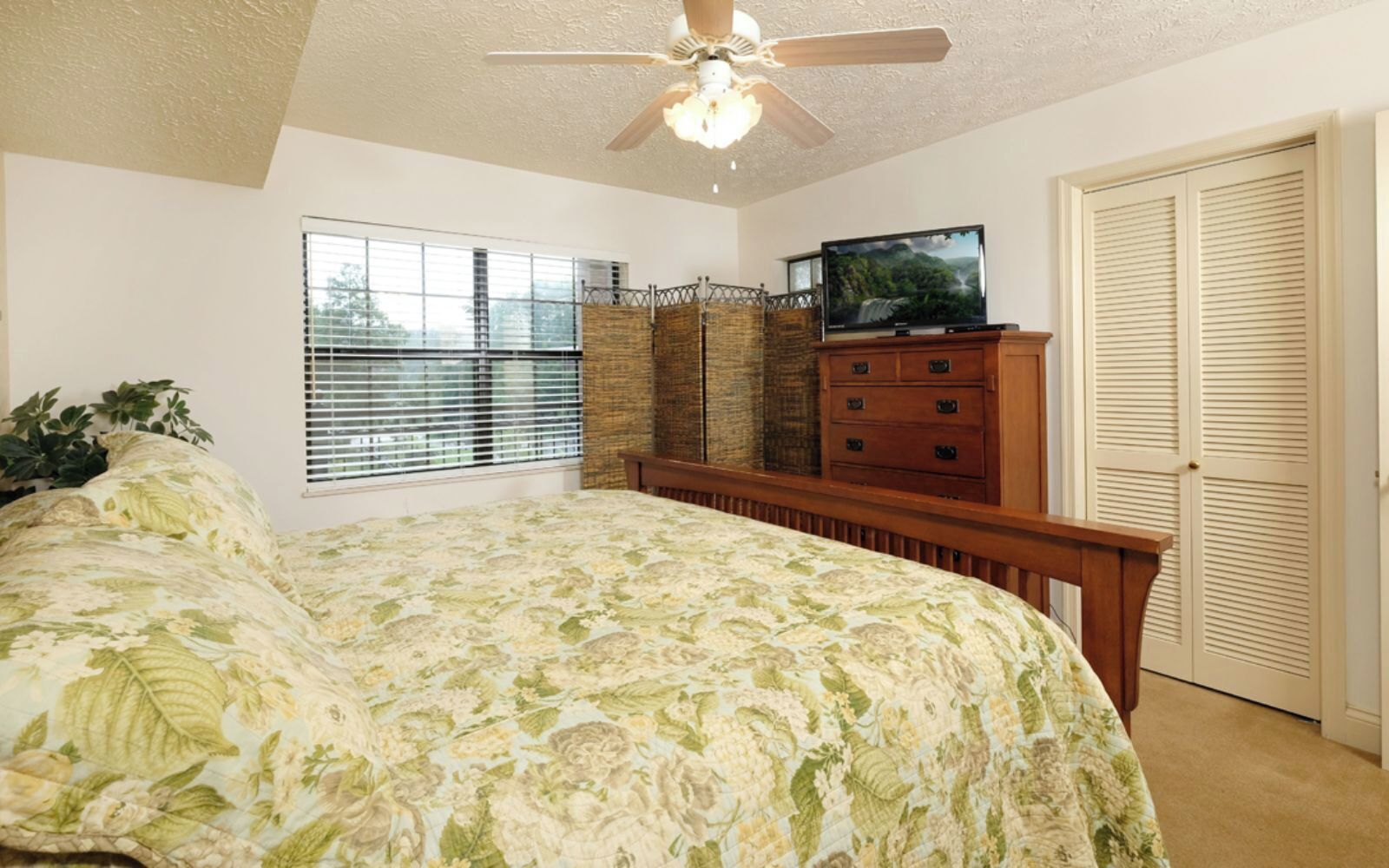 A bedroom with a bed, a television, a wardrobe, a ceiling fan, and a window with blinds.