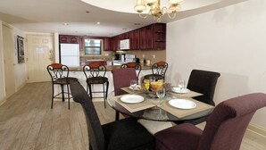 A dining area with a round glass table, six chairs, and a chandelier. A kitchen is visible in the background with wooden cabinets and a refrigerator. - Creek View GH403 - Sip coffee on the balcony with the sound of the creek! (Gatlinburg)