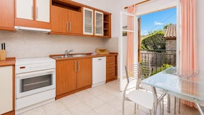 Una cucina compatta con mobili in legno, un piano cottura bianco e una lavastoviglie. Troviamo un tavolo in vetro con sedie all'esterno, vicino a un balcone che offre vista sul verde.