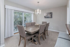 Uma sala de jantar com uma mesa grande, quatro cadeiras, um lustre, um quadro na parede e uma vista pela janela.