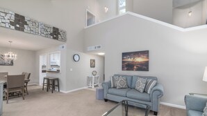 A spacious living area with a high ceiling, a dining table with chairs, a sofa with patterned cushions, and a staircase leading to an upper level.
