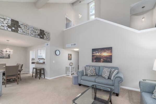 A spacious living area with a high ceiling, a dining table with chairs, a sofa with patterned cushions, and a staircase leading to an upper level.
