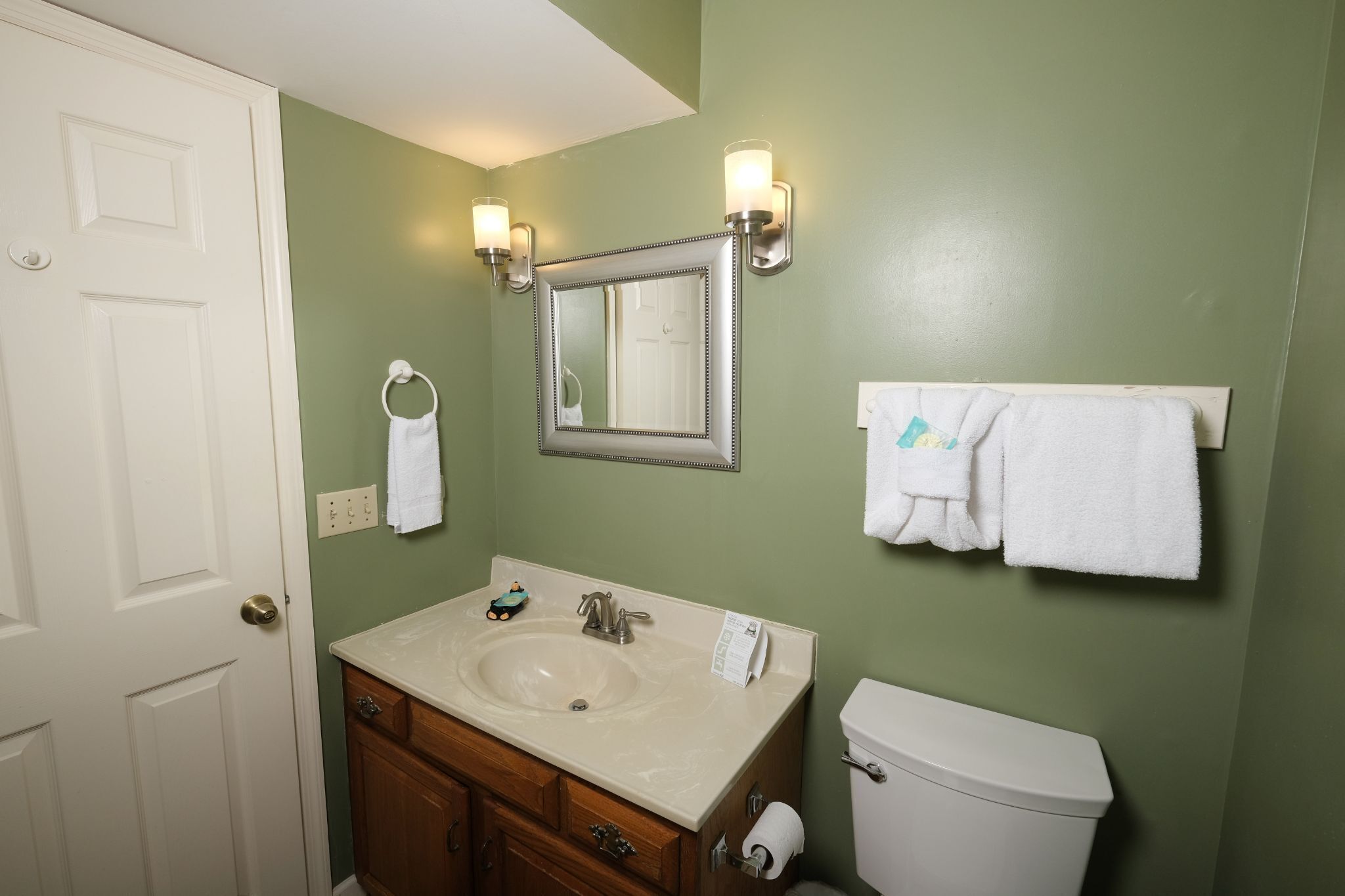 Une salle de bain avec un lavabo, un miroir et des toilettes. Des serviettes sont accrochées au mur et il y a une porte avec une poignée.