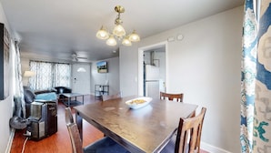 A dining area with a wooden table, chairs, and a bowl of fruit. There is a sofa and a television in the background.