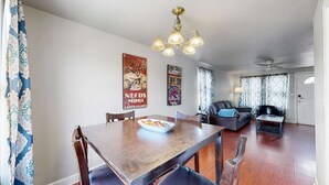 A dining area with a wooden table, chairs, and a bowl of snacks. There are two posters on the wall, a ceiling fan, and a door leading to another room.