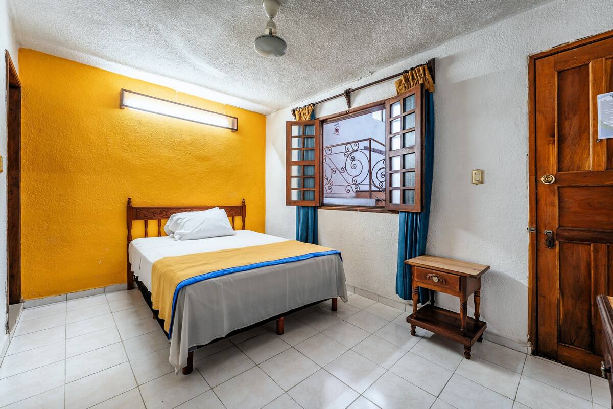 A bedroom with a bed, a nightstand, a wooden door, and a window with curtains.